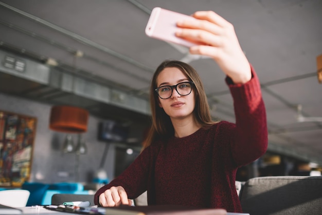 Foto blogger femminile che tiene il cellulare in mano guardando la fotocamera del cellulare e scattando foto di se stessa