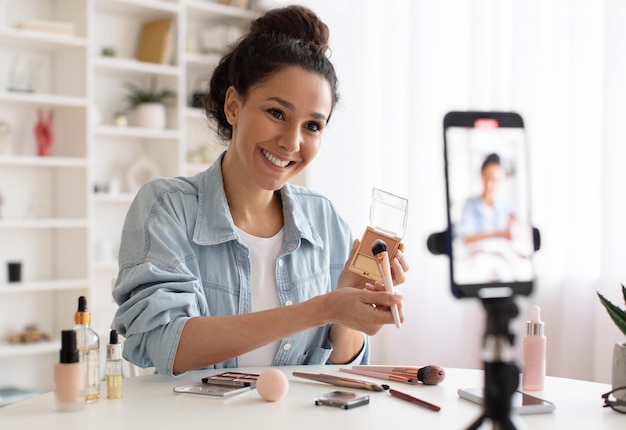 Female blogger showing makeup product to smartphone making video indoor