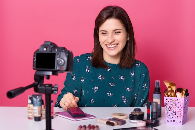 Female blogger sits at table with lots cosmetics products, records tutorial video