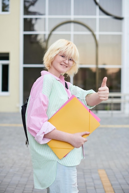 Female blonde student showing cool gesture and standing in front of university campus