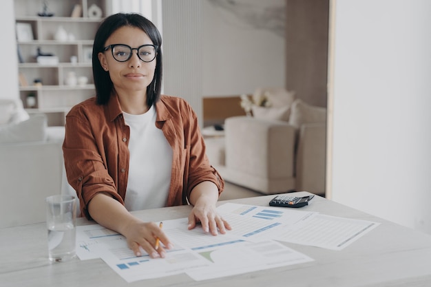 Female bookkeeper freelancer in glasses sitting at desk looking at camera while working from home