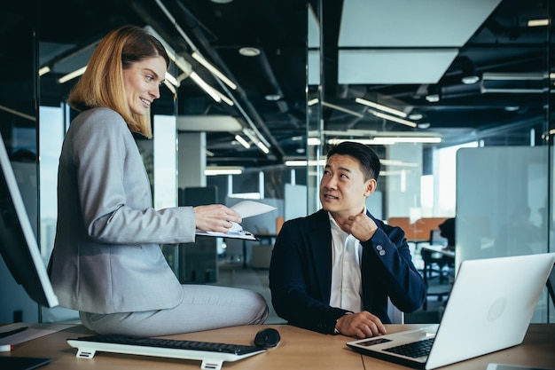 Female boss flirts with male employee businesswoman sitting at desk and smiling then Asian