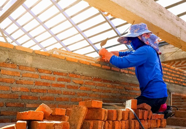 Female bricklayer on wooden platform using trowel to build brick wall in house construction site