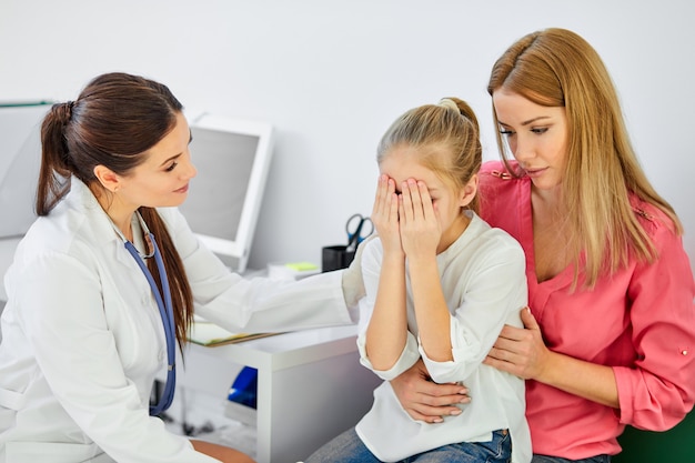 Female calms down the girl patient frightened by medical examination check-up, kid girl sits with mother, closing face with hands