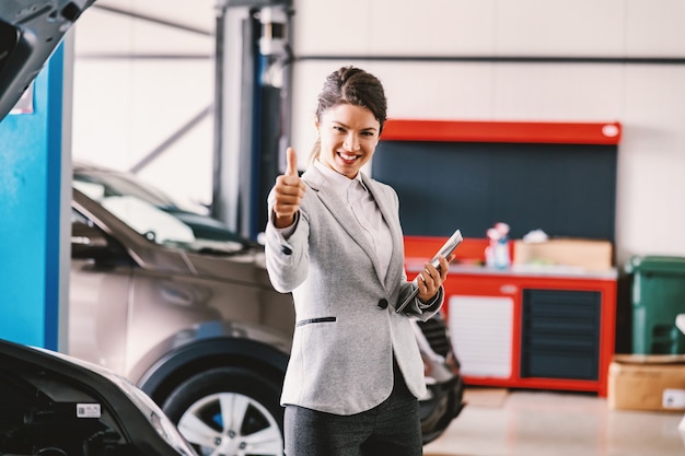 Female car seller standing in garage of car salon and showing thumbs up. Car is all set and repaired.