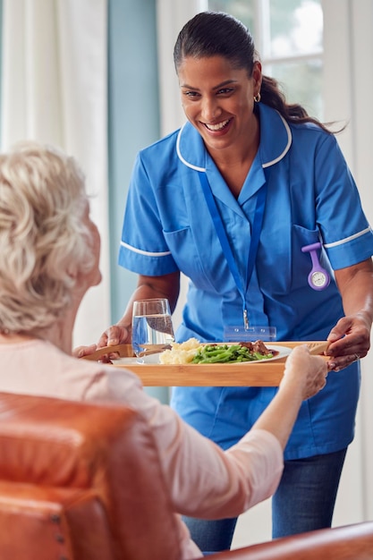 Female Care Worker In Uniform Bringing Meal On Tray To Senior Woman Sitting In Lounge At Home