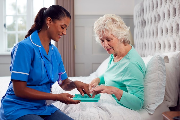 Female care worker in uniform helping senior woman at home in\
bed with medication