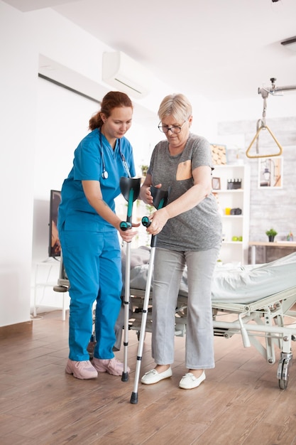 Female caregiver helping elderly age woman in nursing home to walk with crutches.