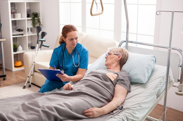 Female caregiver with cliboard talking with sick woman in nursing home.