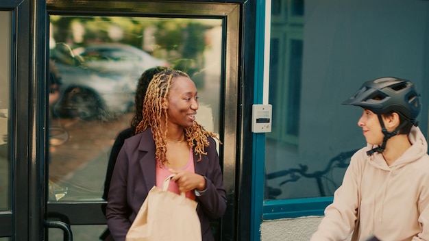 Female carrier giving fastfood package order to woman at office building front door, express delivery. Female client receiving restaurant food in paperbag from courier. Handheld shot.