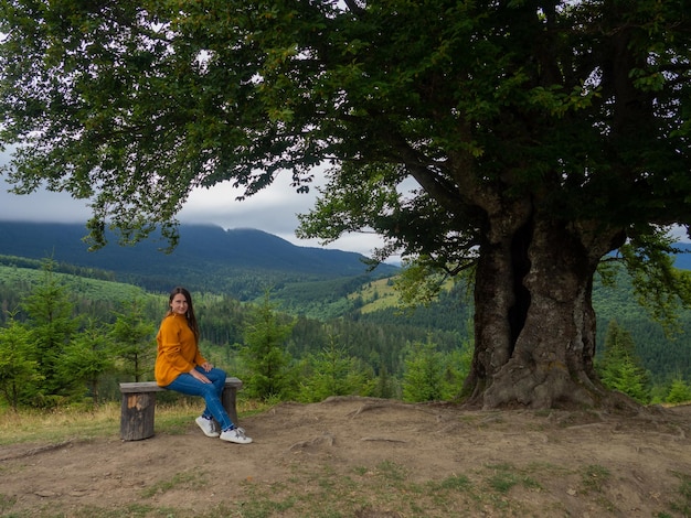 Female in casual clothes sits under large tree with forest background