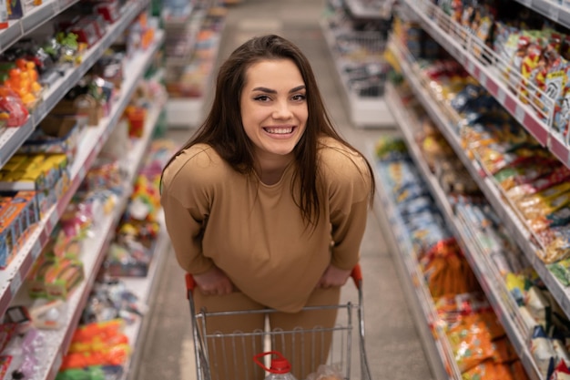 Femmina caucasica cliente in posa con carrello acquisto di cibo in supermercato guardando la fotocamera bruna scegliendo prodotti nel negozio