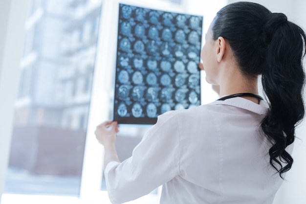 Female charming smart oncologist working at the clinic while examining brain roentgen image and expressing positivity