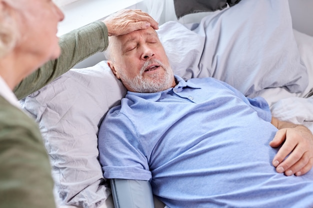 ベッドに横になっている年配の男性の発熱温度をチェックする女性。成熟した夫は、妻が額に触れて熱をチェックしている間、インフルエンザの症状を感じています。自宅で
