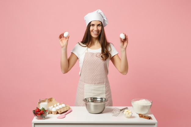 Foto donna chef cuoco pasticcere o fornaio in grembiule t-shirt bianca, cappello da chef toque che cucina torta o cupcake a tavola tenere uova isolate su sfondo rosa pastello in studio. mock up copia spazio concetto di cibo.