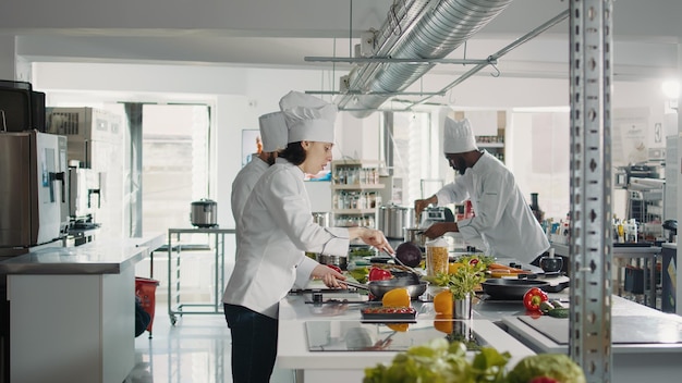 Female chef cooking gastronomy dish with vegetables on stove, making culinary recipe with bell pepper. Woman in uniform preparing food ingredients to cook delicious restaurant meal.