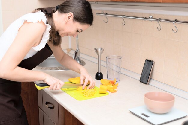 Женщина-шеф-повар режет свежее манго для приготовления домашнего вкусного смузи или сока из манго, используя онлайн-рецепт со смартфона Избирательный фокус Процесс приготовления летнего напитка дома здоровый образ жизни
