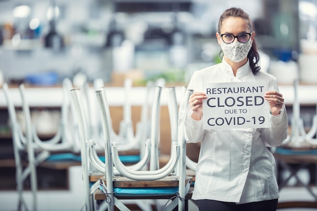 Female chef wearing face mask holds a sign saing restaurant closed due to Covid19