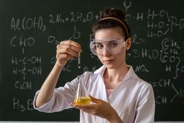 Female chemist doing experiment and looking at colored liquids
in flask, against chalkboard at class