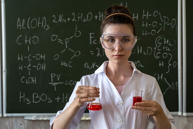 Female chemist doing experiment and looking at colored liquids in flask, against chalkboard at class