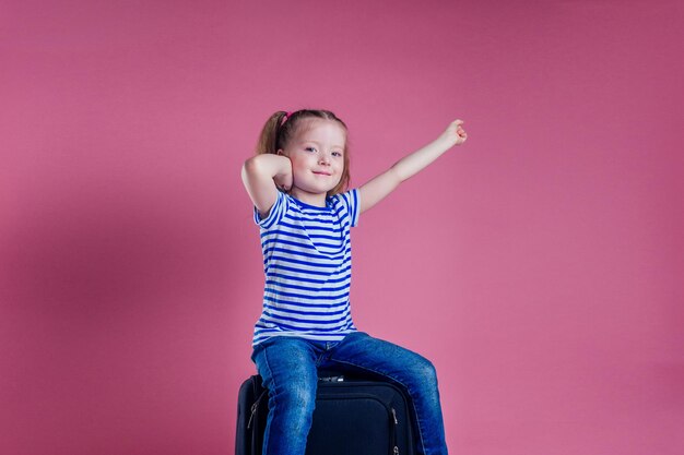 Female child striped blue white t-shirt sitting on a large travel suitcase dream flies rest on the sea shore pink background studio