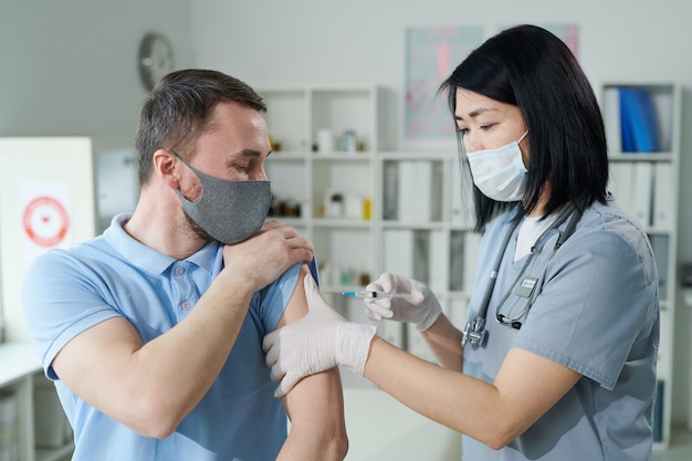 female clinician or nurse of asian ethnicity in uniform and protective mask making injection to young man sitting in front of her in medical office