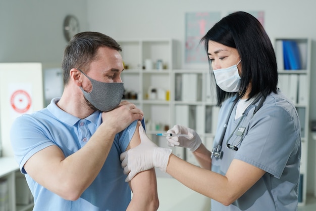 Female clinician or nurse of Asian ethnicity in uniform and protective mask making injection to young man sitting in front of her in medical office