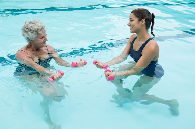 Female coach assisting senior woman in lifting dumbbells in swimming pool