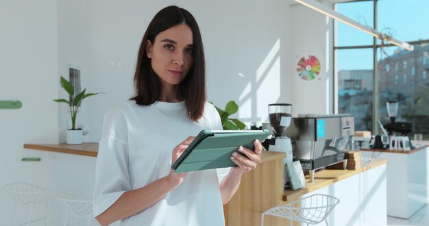 Female coffee shop owner standing in front of counter using digital tablet computer inside modern cafeteria Woman waitress or barista employee of coffeeshop holding modern device