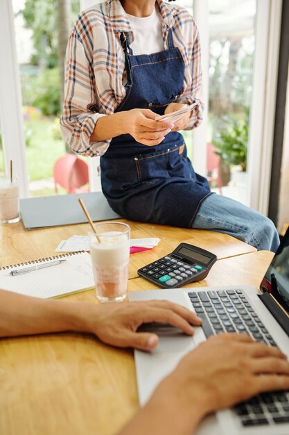 Photo female coffeeshop barista with bills in hands sitting on desk of her boss working on laptop