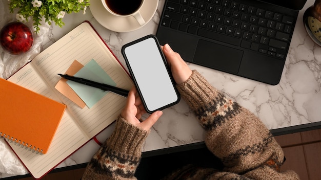 Female college student in cozy sweater using smartphone over her modern workspace. top view