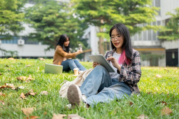 A female college student is studying on her digital tablet while sitting on grass in a park
