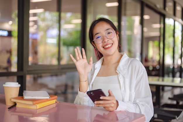 A female college student sits at a table in a campus cafeteria smiling and waving at the camera