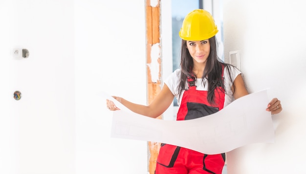 Female construction supervisor studies building plans in the red overall and yellow safety helmet.