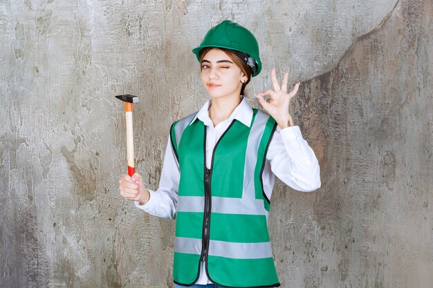 Female construction worker in green helmet holding hammer and giving ok sign