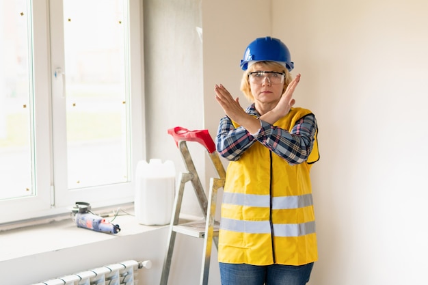 A female construction worker in a new building crossed her arms . A middle-aged woman in a helmet makes repairs in the apartment.