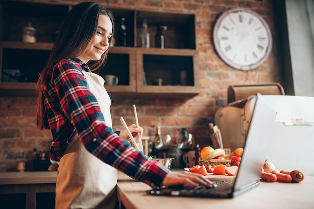 Female cook in apron looks at a recipe in laptop, vegetable salad cooking on the kitchen. Fresh diet food preparation. Wife prepares romantic dinner for her husband