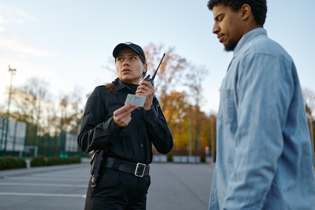 Female cop in black uniform checking male passerby ID document talking to walkie-talkie