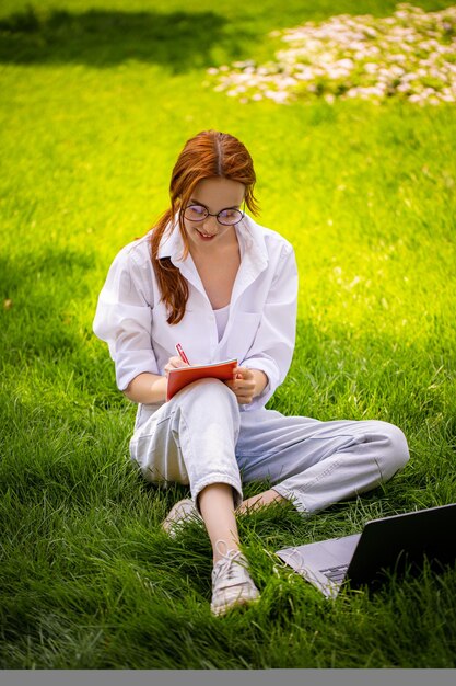 女性のコピーライターは、公園の芝生に座ってノートパソコンのPCモバイルオフィスを使用してメモを作成します