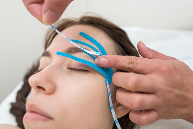 Female cosmetician doing the taping of the face of the patient lying on the desk in the officeSpecialist performing kinesio taping application on girl's face