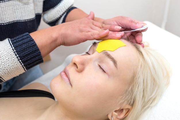Female cosmetician doing the taping of the face of the patient lying on the desk in the officeSpecialist performing kinesio taping application on girl's face