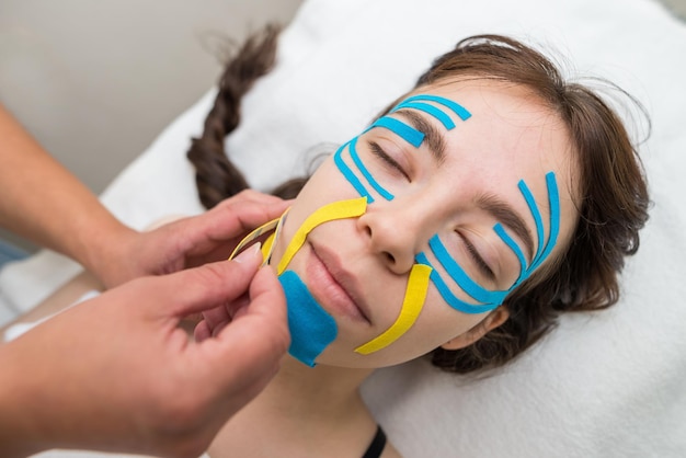 Female cosmetician doing the taping of the face of the patient lying on the desk in the officeSpecialist performing kinesio taping application on girl's face