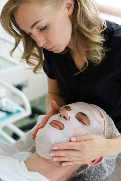 Foto cosmetologo femminile che applica una maschera in tessuto sul viso dei clienti nella clinica di bellezza