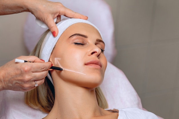 Female cosmetologist drawing an arrow on a woman's face with a white marker to prepare for the procedure.