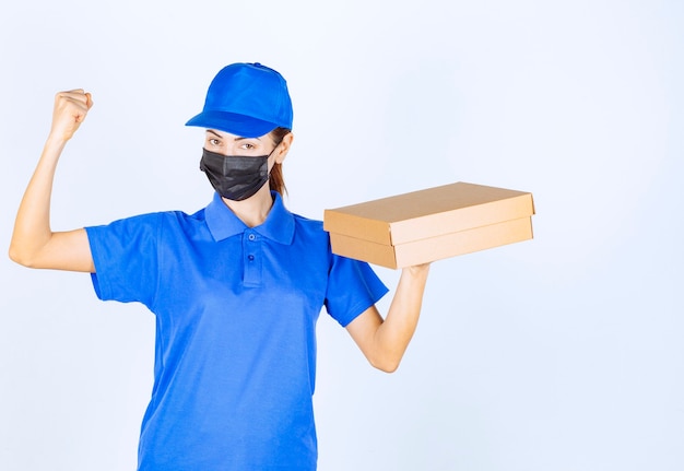 Female courier in blue uniform and face mask holding a cardboard box and showing her fist. 