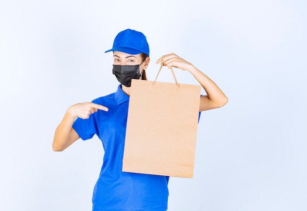 Female courier in blue uniform and face mask holding a cardboard shopping bag. 