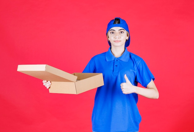 Female courier in blue uniform holding a cardboard takeaway box and showing enjoyment hand sign. 