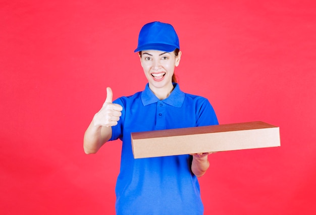 Female courier in blue uniform holding a cardboard takeaway pizza box and showing enjoyment sign. 