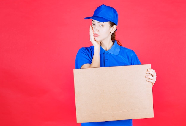 Female courier in blue uniform holding a cardboard takeaway pizza box and thinking or looking confused. 