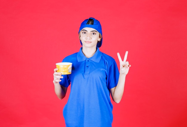 Female courier in blue uniform holding a yellow noodles cup and showing satisfaction sign. 